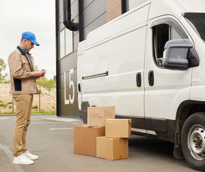 young man uniform using his mobile phone while standing near van packages outdoors near warehouse
